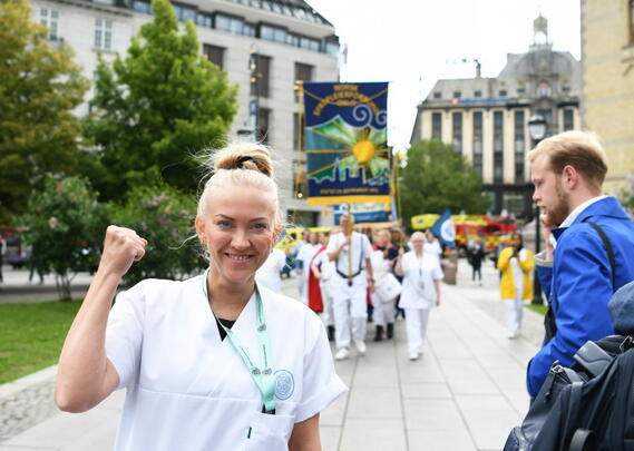 Lill Sverresdatter Larsen i hvit sykepleieruniform smiler til kamera og hever knyttneven. I bakgrunnen ses NSFs fane og sykepleiere i uniform.